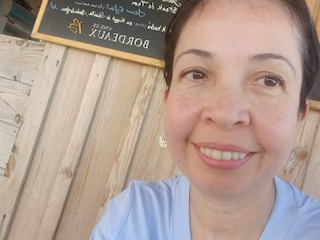 A close-up of Dea smiling at the camera, wearing a light blue t-shirt and with a wooden panel behind her.